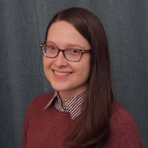 A person with straight, long brown hair is wearing a maroon sweater over a collared shirt with black and white stripes. The background is a textured gray curtain.